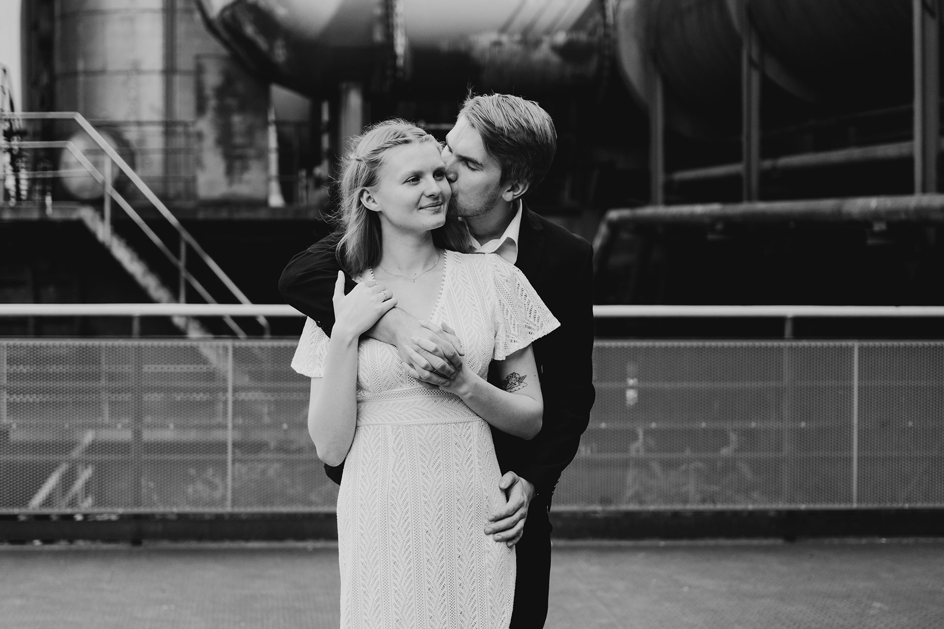 Romantisches Schwarzweiß-Foto von Isabelle und Mark im Landschaftspark Duisburg: Liebevoller Kuss auf die Wange vor malerischer Kulisse - Fotograf: Sugata Tyler